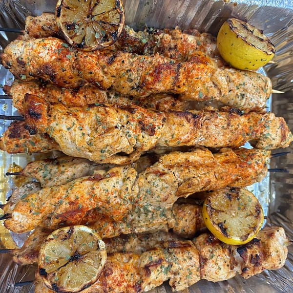 Grilled Greek chicken souvlaki skewers with charred lemon halves in a foil container.
