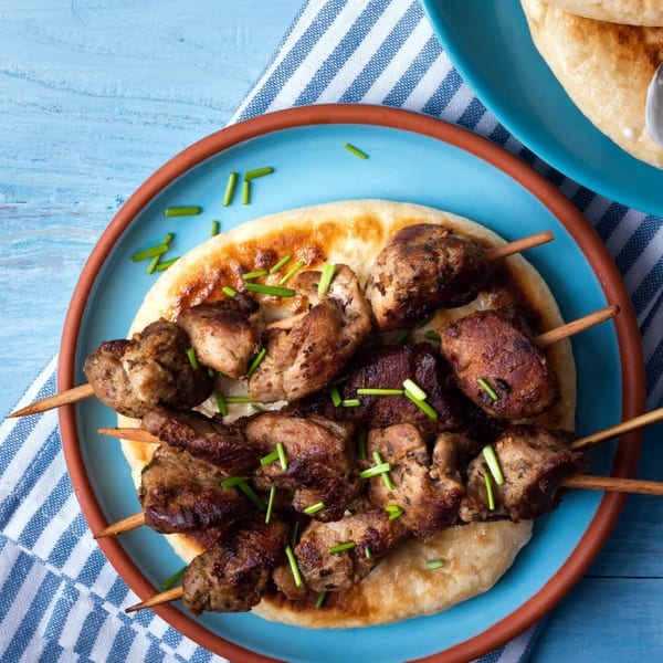 Four Greek Guys Souvlaki skewers with grilled meat on pita bread, garnished with chives on a blue plate.